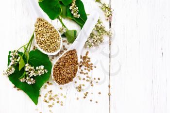 Brown and green buckwheat groats in two spoons with leaves and flowers on the background of a light wooden board from above