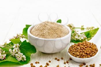 Brown buckwheat flour in a bowl, brown groats in a spoon, buckwheat flowers and leaves on white wooden board background