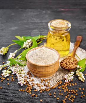 Brown buckwheat flour in a bowl, brown groats in a spoon, buckwheat flowers and leaves, oil in glass jar on burlap on black wooden board background
