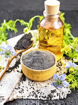Flour of black caraway in a bowl, seeds in a spoon burlap, oil in bottle and twigs Nigella sativa with blue flowers and green leaves on black wooden board background