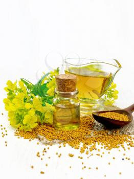 Mustard oil in a glass bottle and gravy boat, grains in a spoon and on a burlap napkin, yellow mustard flowers on background of light wooden board