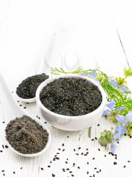 Black cumin seeds in a bowl, flour and seeds in spoons, kalingi sprigs with blue flowers and leaves on a light wooden board background