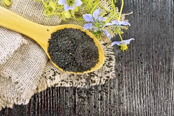 Black cumin seeds in a spoon on sacking, kalingi twigs with blue flowers and green leaves on a wooden board background from above