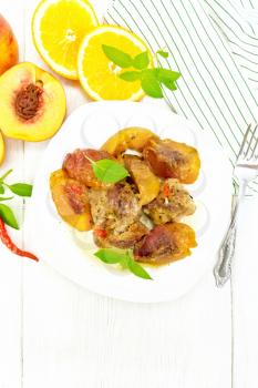Turkey stewed with peaches, fresh hot pepper and orange sauce, basil leaves in a plate, napkin, fruits on light wooden board top background