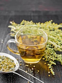 Gray wormwood herbal tea in a glass cup, fresh flowers and metal strainer with dried flowers sagebrush on dark wooden board background
