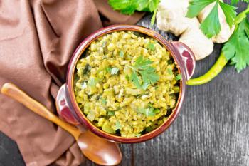 Indian national dish kichari made of mung bean, rice, celery, spinach, hot pepper and spices in a bowl on a towel, ginger and spoon on wooden board background from above