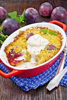 Crumble with plum and ice cream in a red brazier with spoons on a towel, plums and mint on wooden board background