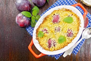 Crumble with plum in a red brazier with spoons on a napkin, plums and mint on wooden board background from above