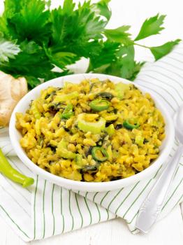 Indian national dish kichari made of mung bean, rice, stalk celery, spinach, hot pepper and spices in a bowl on a striped napkin, ginger on wooden board background