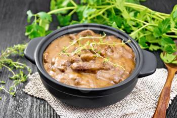 Beef goulash in tomato sauce with thyme sprigs in a pan on burlap, spoon, parsley on black wooden board background