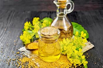 Mustard oil in a glass jar and decanter, mustard grains on a burlap napkin, flowers and leaves on dark wooden board background