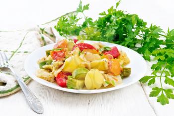 Vegetable ragout with zucchini, cabbage, potatoes, tomatoes and bell peppers in creamy sauce in plate, kitchen towel, parsley and a fork on the background of light wooden board