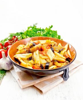 Penne pasta with eggplant and tomatoes in a bowl on kitchen towel, fork, garlic and parsley on a wooden plank background