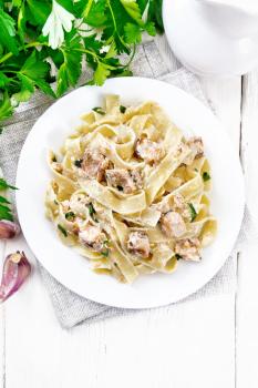 Tagliatelle pasta with salmon, cream, garlic and herbs in a plate on a napkin, fork, parsley and basil on wooden board background from above