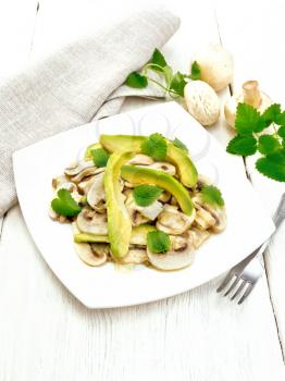 Salad of avocado and raw champignons, seasoned with lemon juice and vegetable oil with mint leaves, a towel and a fork on the background of a wooden board