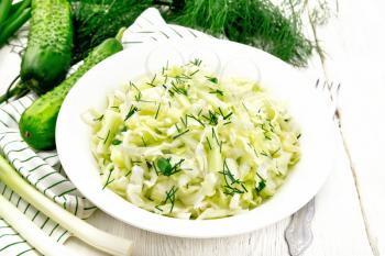 Salad of fresh cabbage, green onions and cucumber with vinegar and vegetable oil dressing in a plate, napkin, dill and fork on white wooden board background