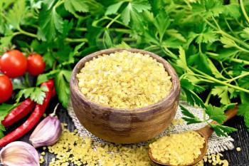 Bulgur groats - steamed wheat grains - in a clay bowl and a spoon on burlap, tomatoes, hot peppers, garlic and parsley on wooden board background