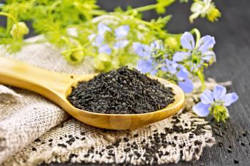 Black cumin seeds in a spoon on sacking, kalingi twigs with blue flowers and green leaves on a wooden board background