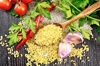 Bulgur groats - steamed wheat grains - in a spoon on sacking, tomatoes, hot peppers, garlic and parsley on a wooden board background from above