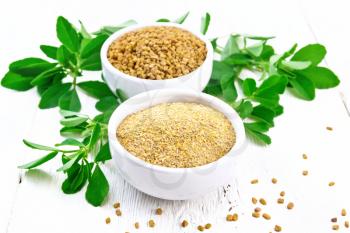 Fenugreek seeds and ground spice in two bowls and on a table with green leaves on background of light wooden board