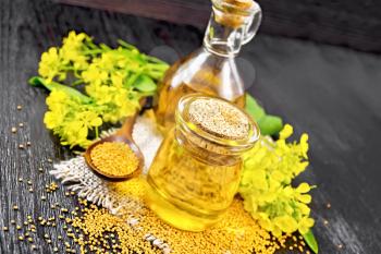Mustard oil in a glass jar and decanter, mustard grains on a burlap napkin, flowers and leaves on black wooden board background