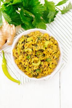 Indian national dish kichari made from mung bean, rice, stalk celery, spinach, hot pepper and spices in a bowl on a napkin, ginger on light wooden board background from above