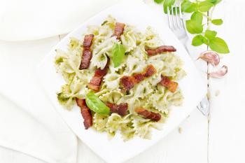 Farfalle pasta with pesto sauce, fried bacon and basil in a plate, garlic, fork and a napkin on light wooden board background from above