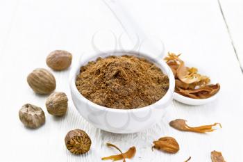 Ground nutmeg in a bowl and dried nutmeg arillus in a spoon, whole nuts on light wooden board background