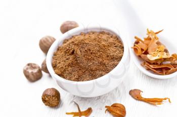 Ground nutmeg in a bowl and dried nutmeg arillus in a spoon, whole nuts on white wooden board background