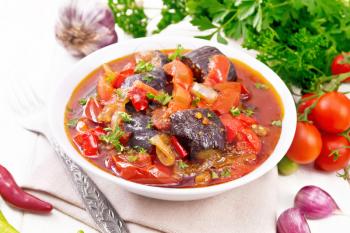 Vegetable ragout with eggplant, tomatoes, bell peppers, onions and spices in a plate on towel, garlic, parsley, hot peppers and a fork on the background of light wooden board
