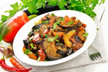 Vegetable ragout with eggplant, tomatoes, sweet and hot peppers, onions, carrots, fried with herbs and spices in plate on a napkin, garlic, parsley on the background of light wooden board