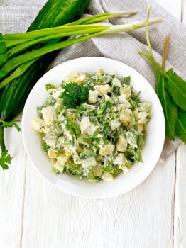 Salad of cucumber, sorrel, boiled potatoes, eggs and herbs, dressed with mayonnaise in a white plate, parsley, green onions and napkin against wooden board on top