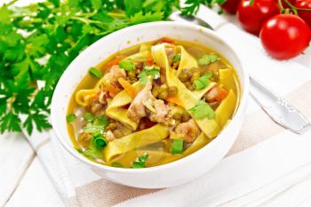 Soup with meat, tomatoes, vegetables, mung bean lentils and noodles in a bowl on a napkin, parsley and spoon on the background of light wooden board
