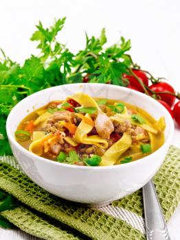 Soup with meat, tomatoes, vegetables, mung bean lentils and noodles in a bowl on a green napkin, parsley and spoon on the background of light wooden board