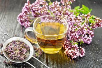 Oregano herbal tea in a glass cup, fresh marjoram flowers and a metal strainer with dried flowers on dark wooden board background