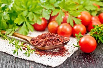 Dried tomato flakes in a spoon on a burlap napkin, fresh small tomatoes, parsley and thyme on wooden board background