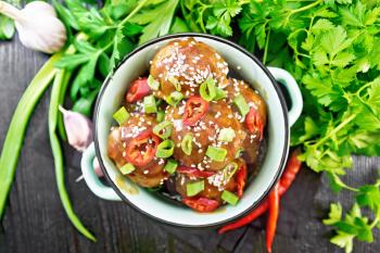 Korean meatballs in sauce of starch, soy sauce, vinegar and apricot jam sprinkled with green onions, hot peppers and sesame seeds in a saucepan on a napkin against wooden board from above