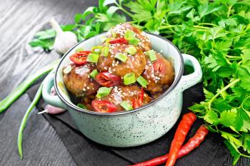 Korean meatballs in sauce of starch, soy sauce, vinegar and apricot jam, sprinkled with green onions, hot peppers and sesame seeds in a saucepan on a towel against wooden board background