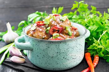 Korean meatballs in sauce of starch, soy sauce, vinegar and apricot jam, sprinkled with green onions, hot peppers and sesame seeds in a saucepan on a napkin against black wooden board