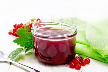Red currant jam in a glass jar, bunches of berries with leaves, a towel and a spoon on wooden board background