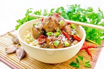 Korean meatballs in sauce of starch, soy sauce, vinegar and apricot jam, sprinkled with green onions, hot peppers and sesame seeds in a bowl on a bamboo napkin on wooden board background