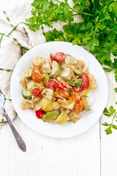 Vegetable ragout with zucchini, cabbage, potatoes, tomatoes and bell peppers in creamy sauce in plate, a towel, parsley and a fork on wooden board background from above