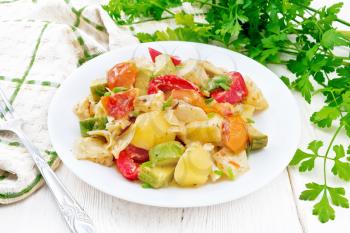 Vegetable ragout with zucchini, cabbage, potatoes, tomatoes and bell peppers in creamy sauce in plate, a towel, parsley and a fork on the background of light wooden board