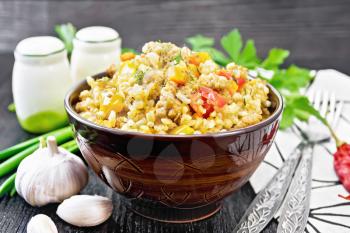 Barley porridge with minced meat, yellow and red bell peppers, garlic and onions in a clay bowl, a towel and parsley on dark wooden board background