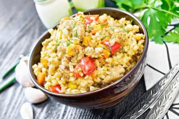 Barley porridge with minced meat, yellow and red bell peppers, garlic and onions in a clay bowl, napkin and parsley on black wooden board background