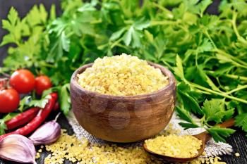 Bulgur groats - steamed wheat grains - in a clay bowl and a spoon on sacking, tomatoes, hot peppers, garlic and parsley on black wooden board background