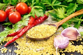 Bulgur groats - steamed wheat grains - in a spoon on sacking, tomatoes, hot peppers, garlic and parsley on a wooden board background