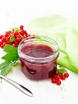 Red currant jam in a glass jar, bunches of berries with leaves, a napkin and a spoon on white wooden board background