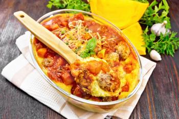 Corn porridge with meatballs, sauce of tomato, garlic and pumpkin, sprinkled with cheese, basil and spoon in a pan on kitchen towel against the background of dark wooden board