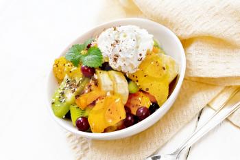 Fruit salad with banana, orange, kiwi, cranberries and baked pumpkin, whipped cream, sprinkled with chocolate and coconut with mint in a bowl on a napkin on white wooden board background